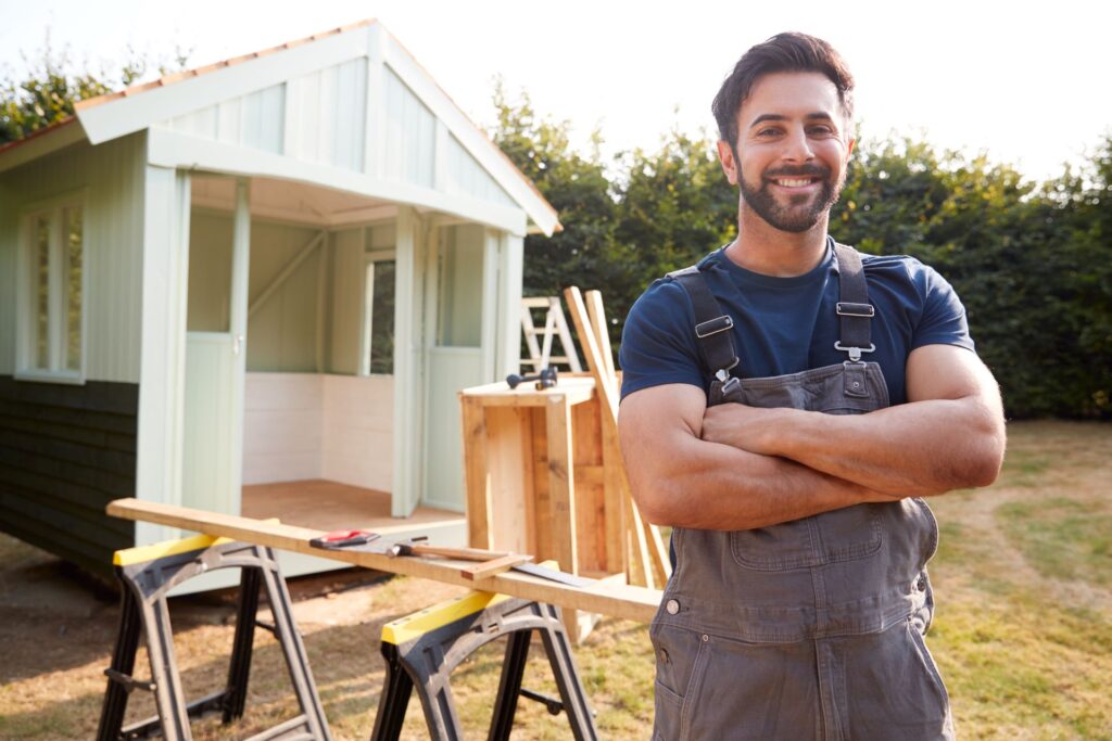 Lächelnder Handwerker mit verschränkten Armen steht vor einem neu renovierten Gartenhaus, vermittelt Tipps zum Armaturen kaufen und effiziente Renovierungstechniken.