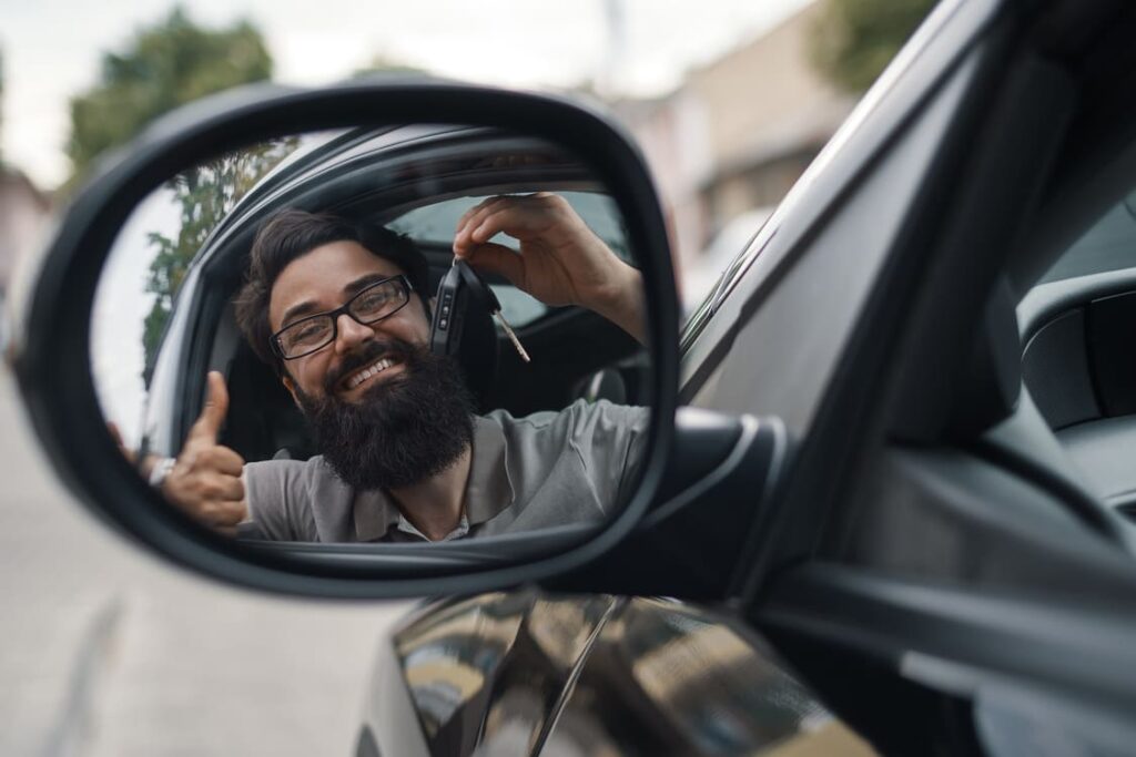 Ein glücklicher Mann mit Brille und Bart zeigt seinen neuen Mercedes Ersatzschlüssel durch den Seitenspiegel seines Autos und hebt den Daumen hoch, nachdem er den Schlüssel nachmachen ließ.