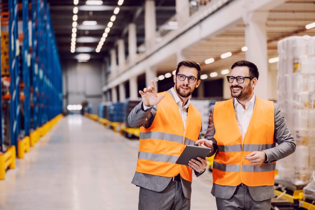 Zwei Logistikmanager in einer Lagerhalle tragen Schutzwesten und diskutieren über Lagerprozesse. Einer hält ein Tablet in der Hand, während der andere auf einen Punkt in der Halle zeigt. Die Szene zeigt eine moderne Lagerumgebung