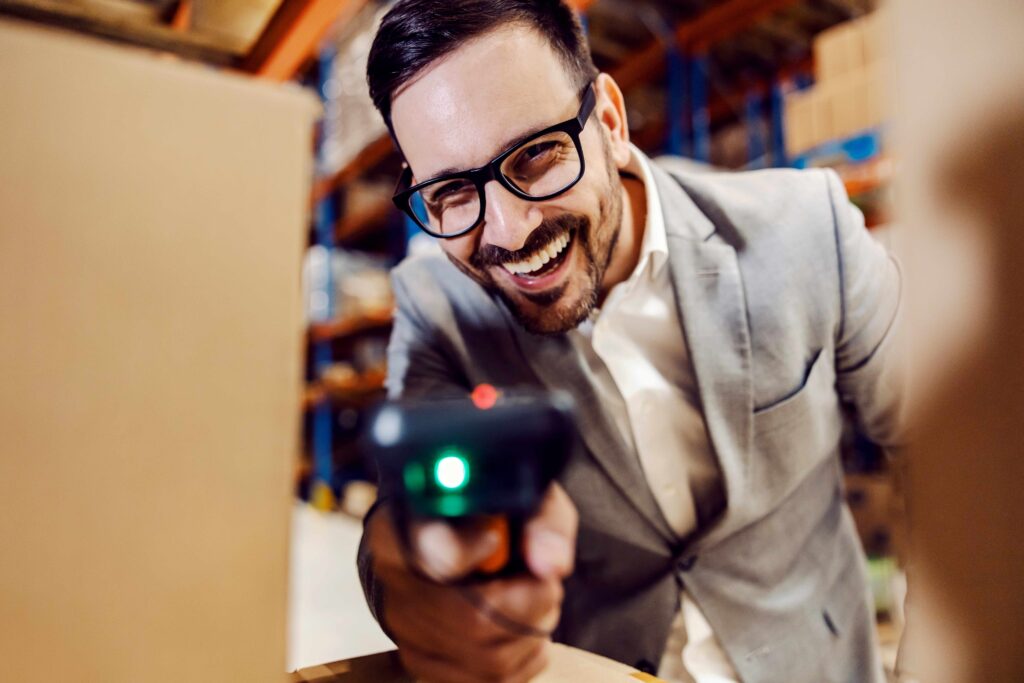 Ein lachender Mann mit Brille scannt Pakete mit einem Barcodescanner in einer Lagerhalle. Der Mann trägt einen grauen Anzug und zeigt Freude bei der Arbeit. Das Bild vermittelt eine positive und effiziente Arbeitsatmosphäre