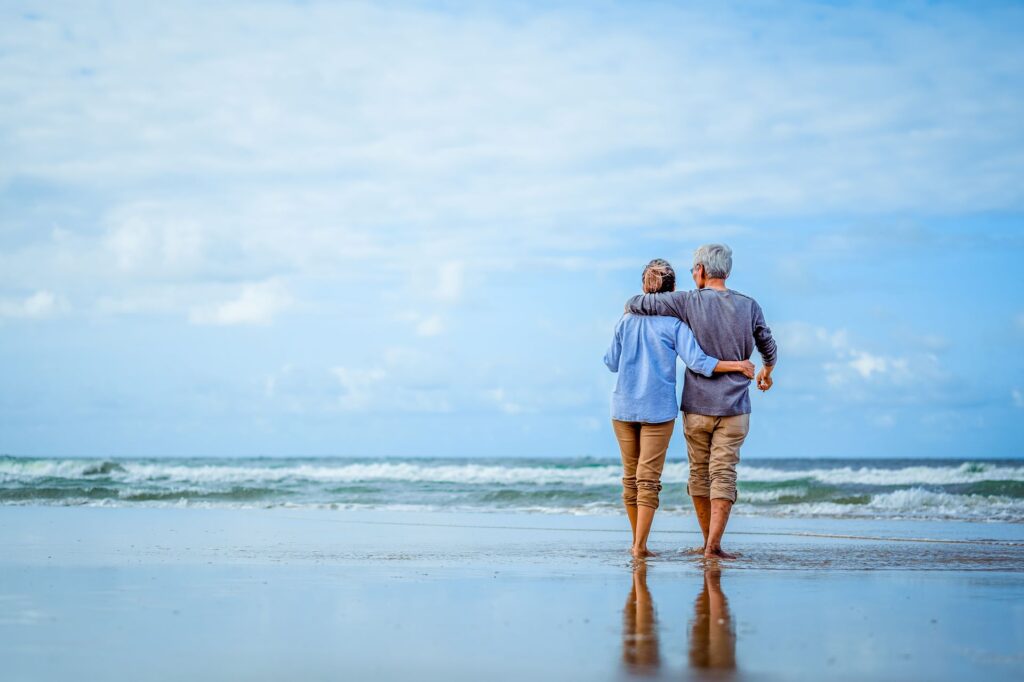 Ein älteres Paar läuft Arm in Arm am Strand entlang und genießt die gemeinsame Zeit. Das Bild symbolisiert Ruhe, Entspannung und eine gesteigerte Lebensqualität im Ruhestand.