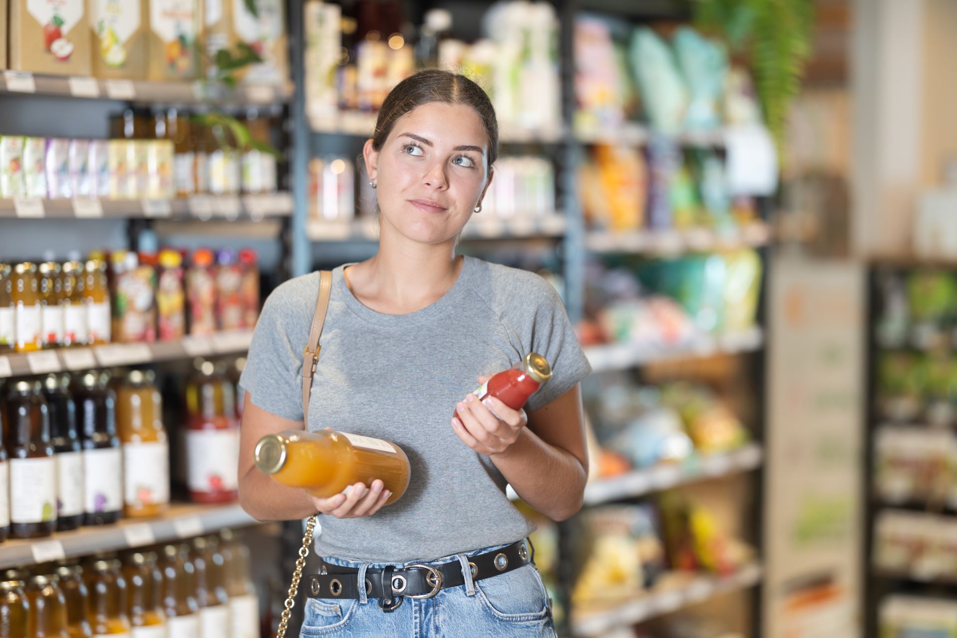 Junge Frau steht nachdenklich vor einem Supermarktregal und hält zwei Flaschen in den Händen, während sie über ihre Kaufentscheidung nachdenkt – Symbolbild für bewussten Einkauf und Preisvergleich.