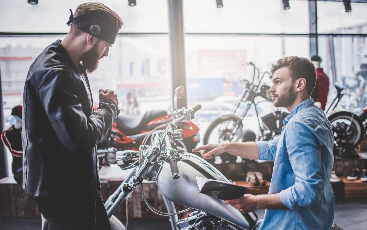 Zwei Männer in einem Motorradgeschäft: Ein Berater erklärt einem Kunden die Vorteile von Zubehör wie der SW Motech Hecktasche, um das Gepäck zu optimieren.