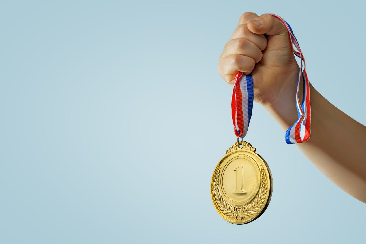 Hand haelt goldene Eins-Medaille mit Band vor hellblauem Hintergrund – Symbol fuer Erfolg und Motivation – Medaille kaufen